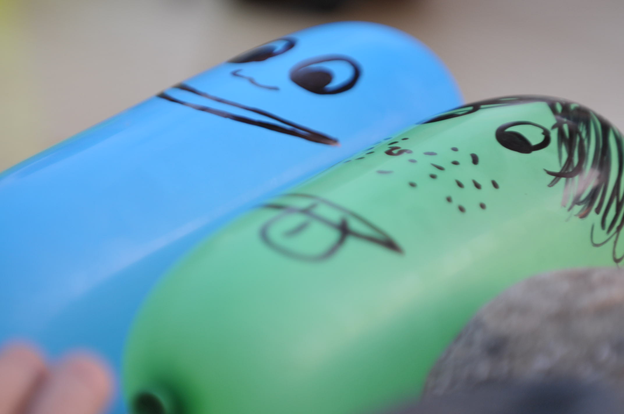 Zwei längliche Ballone. Der blaue hat ein lächelndes Gesicht; der grüne streckt die Zunge heraus und hat Sommersprossen.