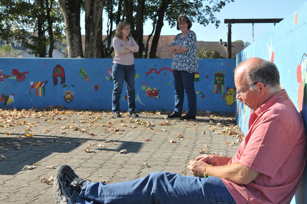 Noldi geniesst im Vordergrund, mit ausgestreckten Beinen sitzend, das sonnige Wetter während der Pause. Im Hintergrund stehen Miryam und Sandra.